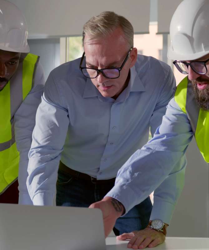 Three men, two of them in full PPE, leant over a desk looking at a laptop.