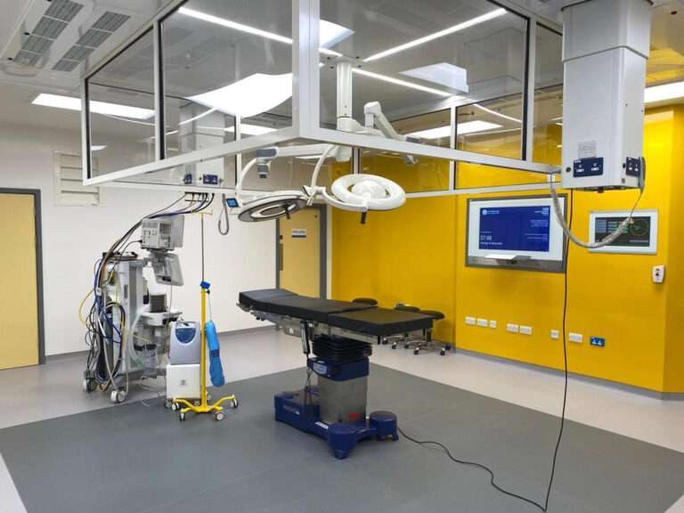 A new operating theatre. The rearmost wall is yellow and features a couple of digital screens. In the foreground there is an operating table with lights over the top, as well as a number of items of medical equipment.