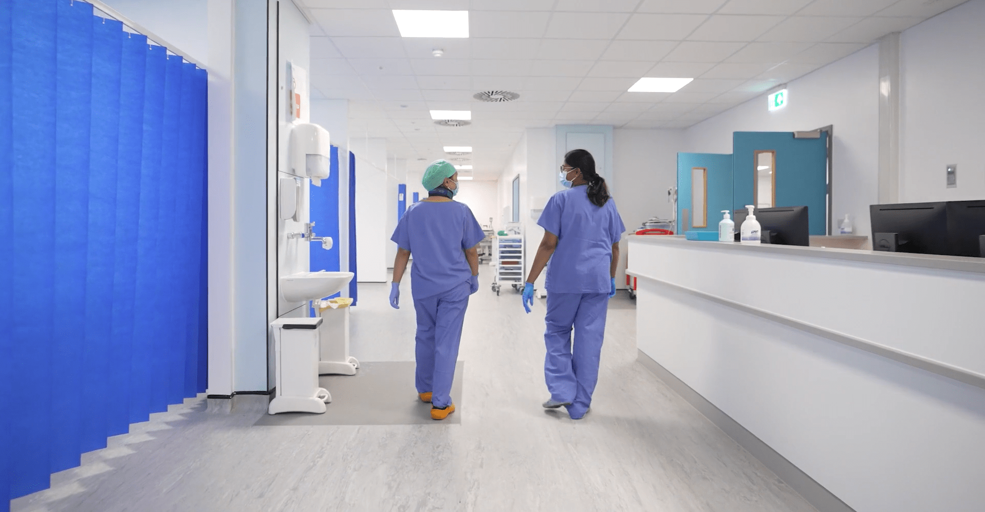 Two clinicians in blue scrubs, walking down a brightly lit, new hospital ward, with bed bays to their left, surrounded by blue curtains, and a nurse's station to the right.