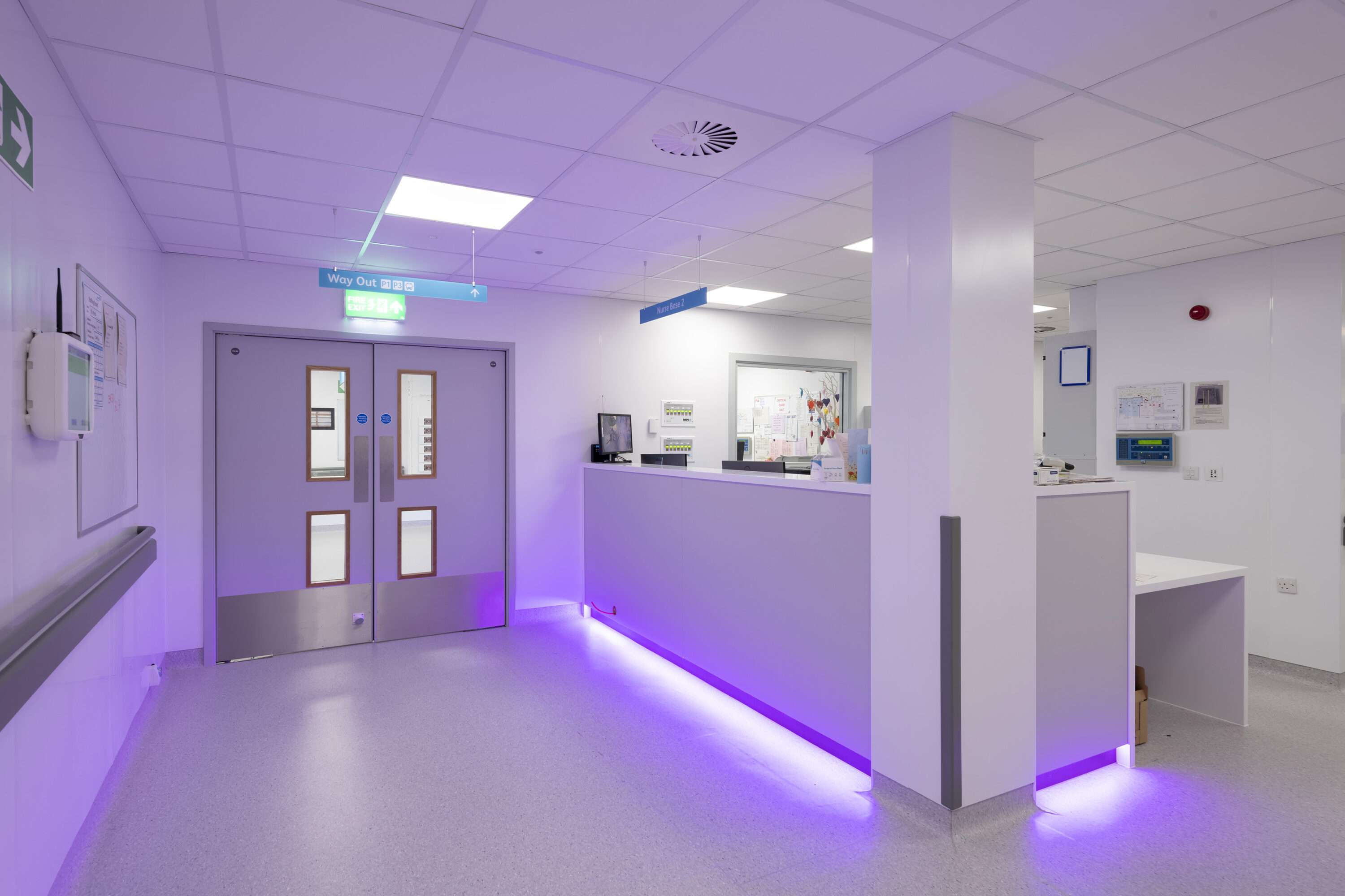 A nurses' station at the side of a hospital corridor. To the left there is a closed double door. The station is to the right. The walls and ceiling are white, the floor is finished in light grey mottled vinyl and the nurses' station features purple ambient lighting.