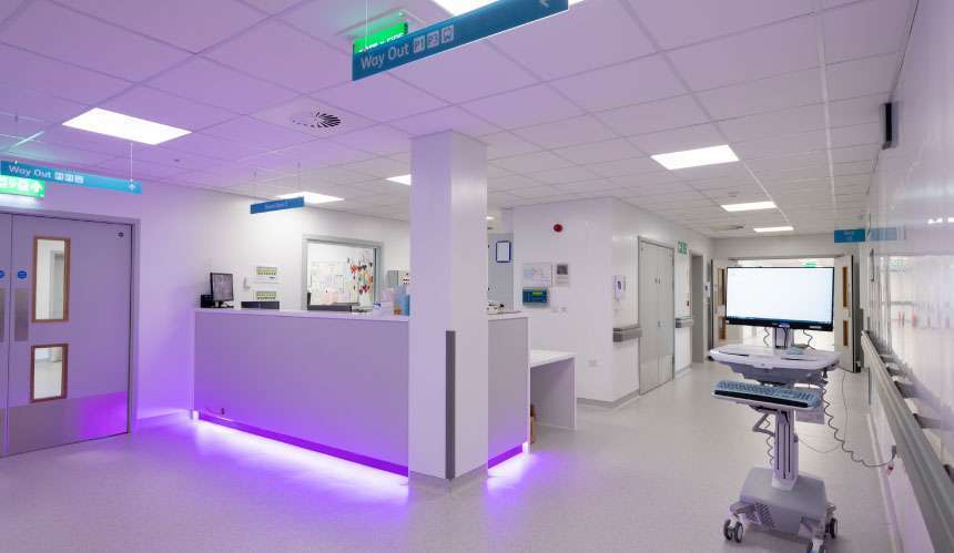 A hospital corridor featuring a nurses' station. The walls are white, there is overhead lighting and the station features purple ambient lighting. To the left is a set of double doors, to the right the corridor continues through another set of doors and there is a trolley containing some medical equipment in the foreground.