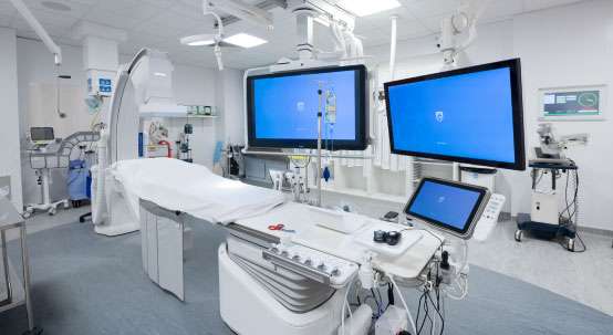 A cardiac catheterisation laboratory. It is white and very light, with medical equipment on three sides and a patient bed with overhead lights, diagnostic screens and equipment in the foreground.