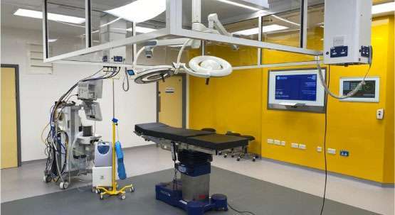 A new operating theatre. The rearmost wall is yellow and features a couple of digital screens. In the foreground there is an operating table with lights over the top, as well as a number of items of medical equipment.