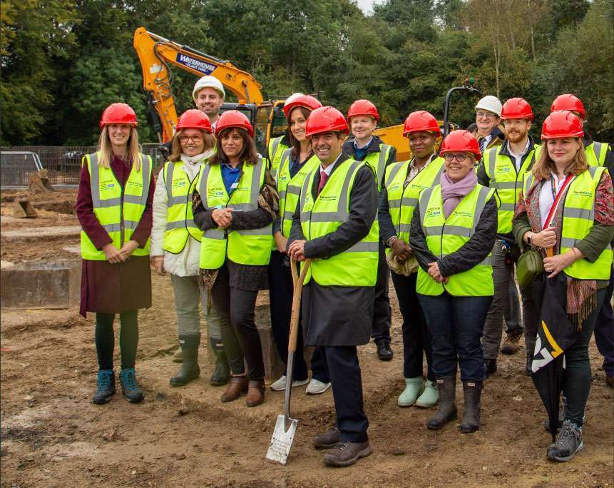 Ground breaking event. A mixed group of people in high-visibility vests and hard hats, standing outside in a building site. The foremost person is holding an engraved ceremonial spade. In the background there is some plant machinery and a screen of trees.