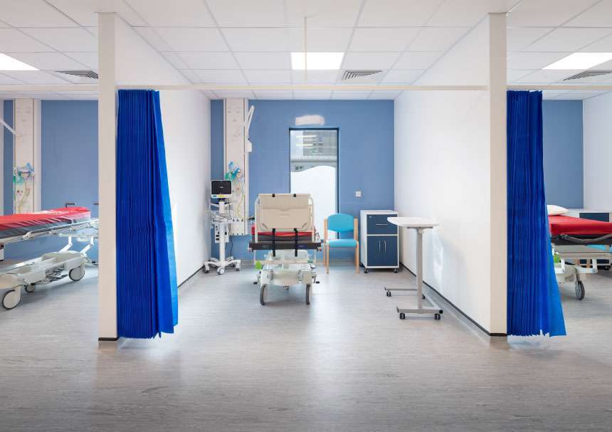 A series of hospital beds in individual bays. The rear wall is a powder blue and the curtains are a darker blue. Each bay has a hospital bed, bedside cabinet and medical equipment in it.