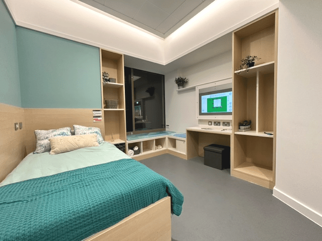 Inside the Bedroom Evolved. The floor is grey, the walls a soft green and all of the wood trim is a light oak colour. There is a digital screen to the right hand side, a bed to the left and small shelves, a window seat and a large window at the far end.