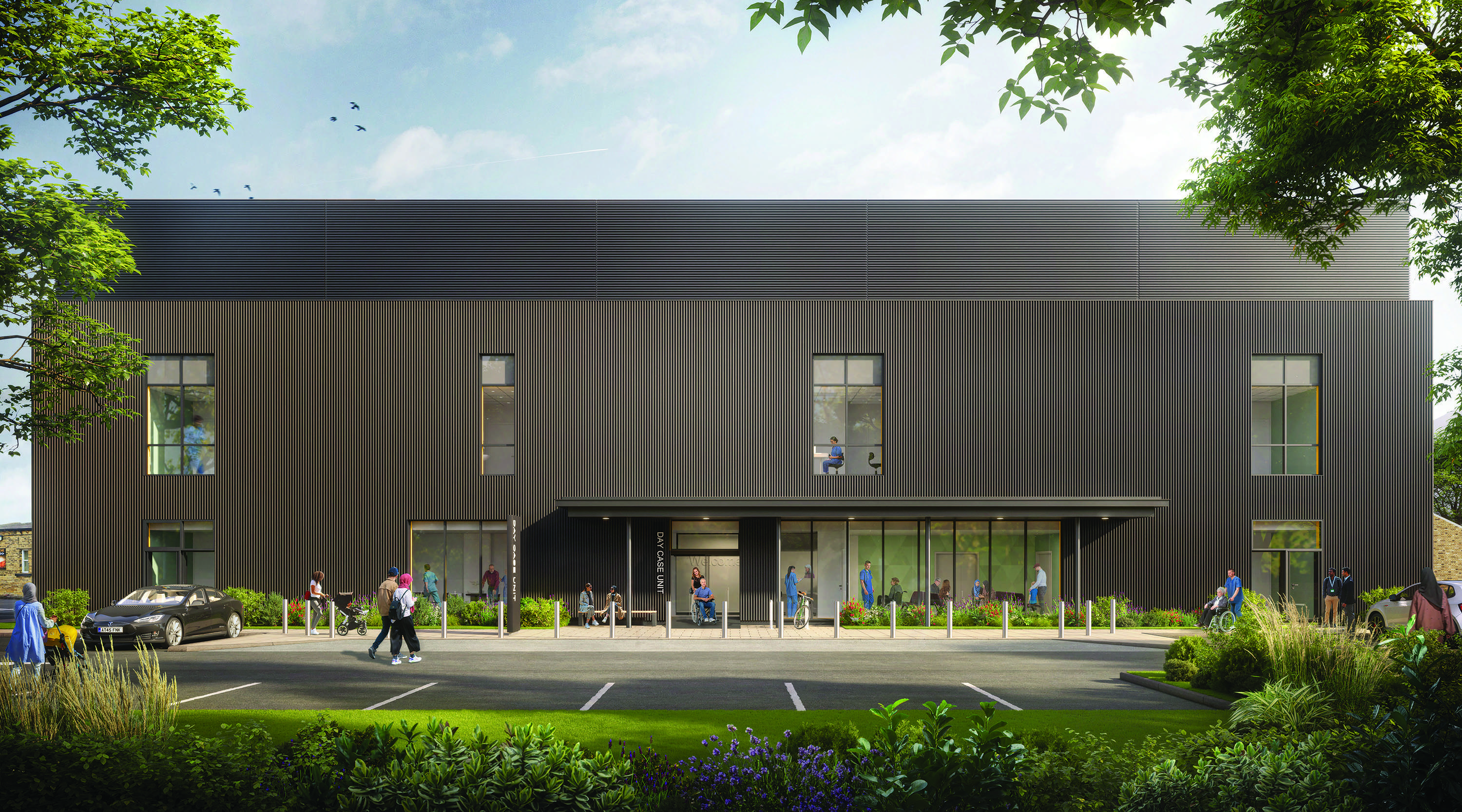 A flat-on elevation of the new Day Case Unit at St Luke's, Bradford. The entrance has a wide canopy over it and a long glass area. There are large windows to the ground and upper floors. The cladding consists of dark, thin strips. At the front of the building there are parking bollards, parking spaces and in the foreground an area of greenery and mature trees. A number of people are inside and outside the building.