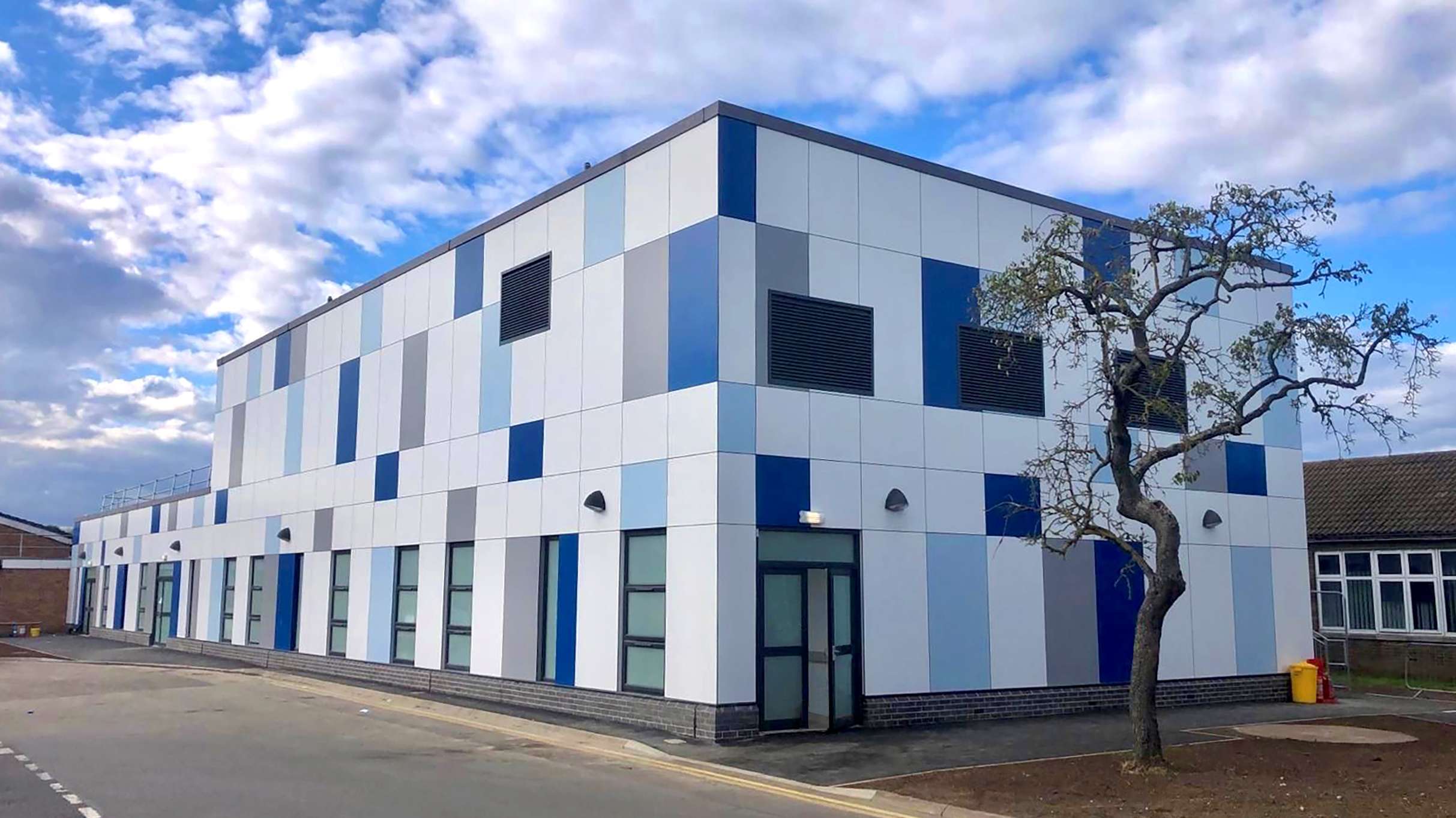 Three-quarter angle photo of the exterior of the Grantham project. The cladding of the building is a patchwork of cladding panels in white, grey and two shades of blue. The ground floor has a number of large windows and a double door to one end. In the foreground there is a large tree which is just beginning to come into leaf.
