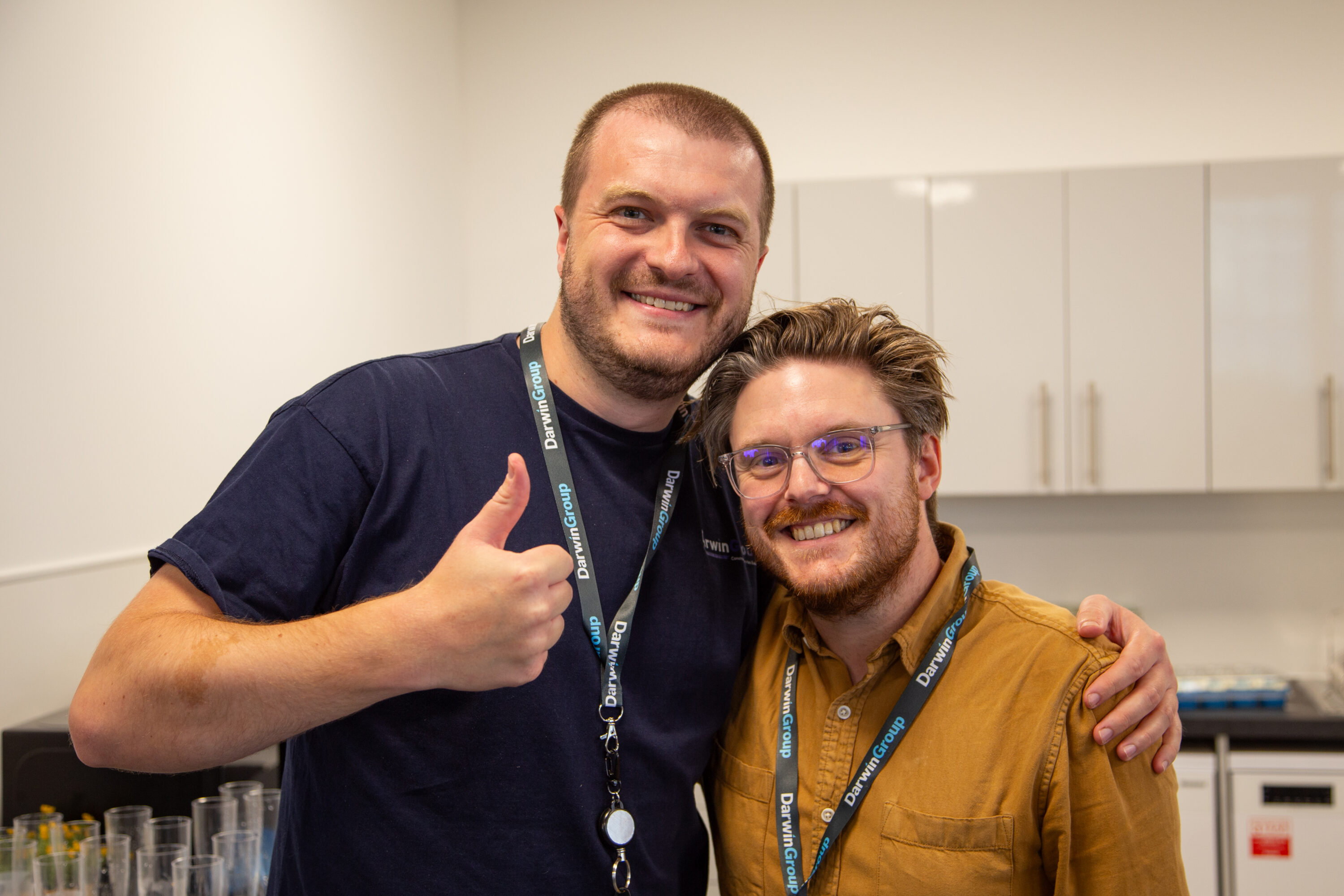 Two men smiling at the camera, one has his thumb up and has his arm around the other man's shoulders.