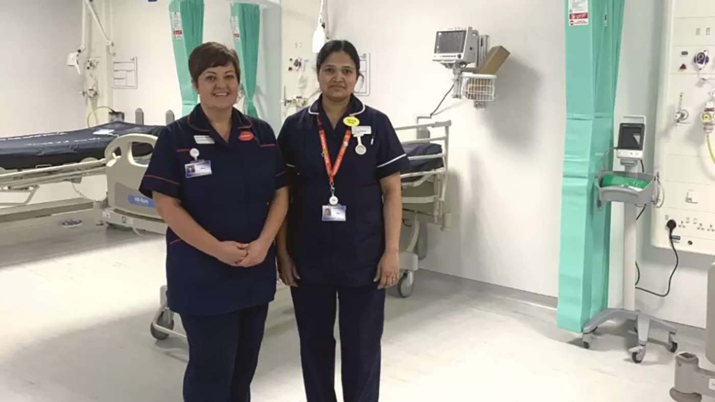 Two clinicians in dark blue uniforms stand in a hospital ward.