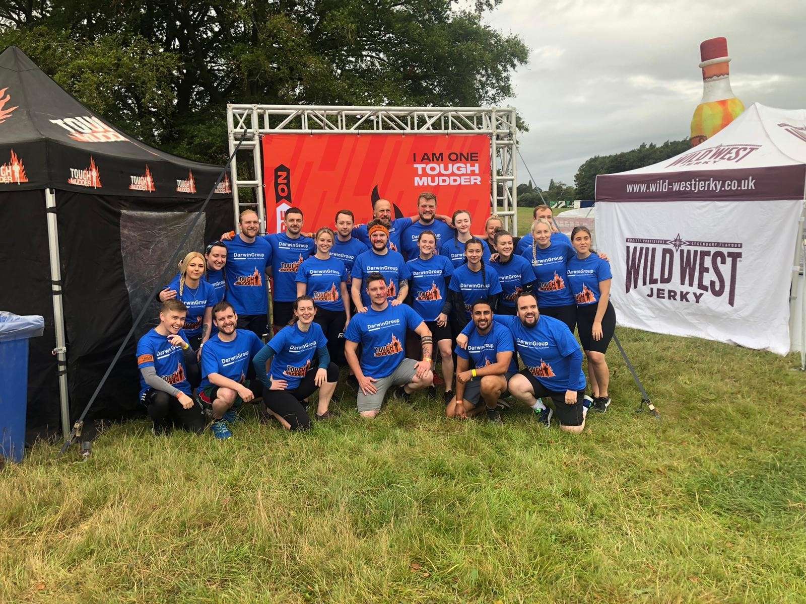 The Darwin Group team in a group photo before they started the Tough Mudder challenge. They're in a grass field with gazebos to either side.