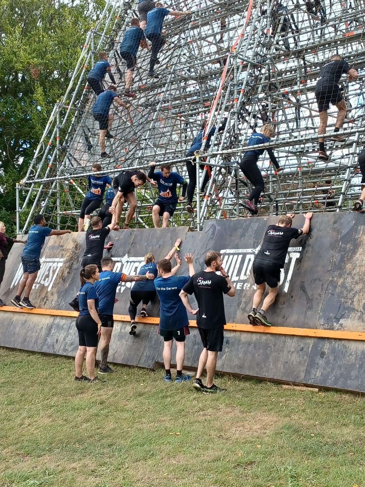 A large climbing obstacle, with lots of people scaling it and helping others to get up and onto it.