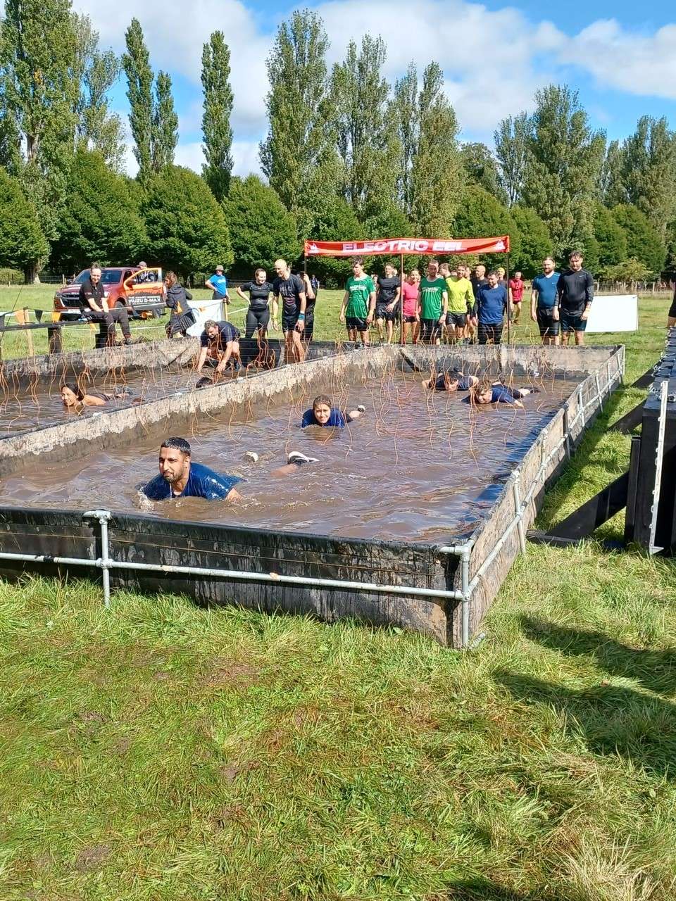 People crawling through a muddy water obstacle, with electric wires hanging down. There are trees and blue sky in the background.