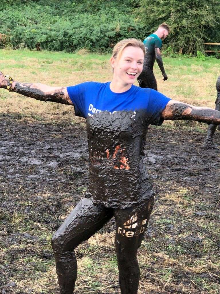 A smiling female runner with her arms stretched out wide. She is covered up to her chest in mud, there is a muddy field in the background