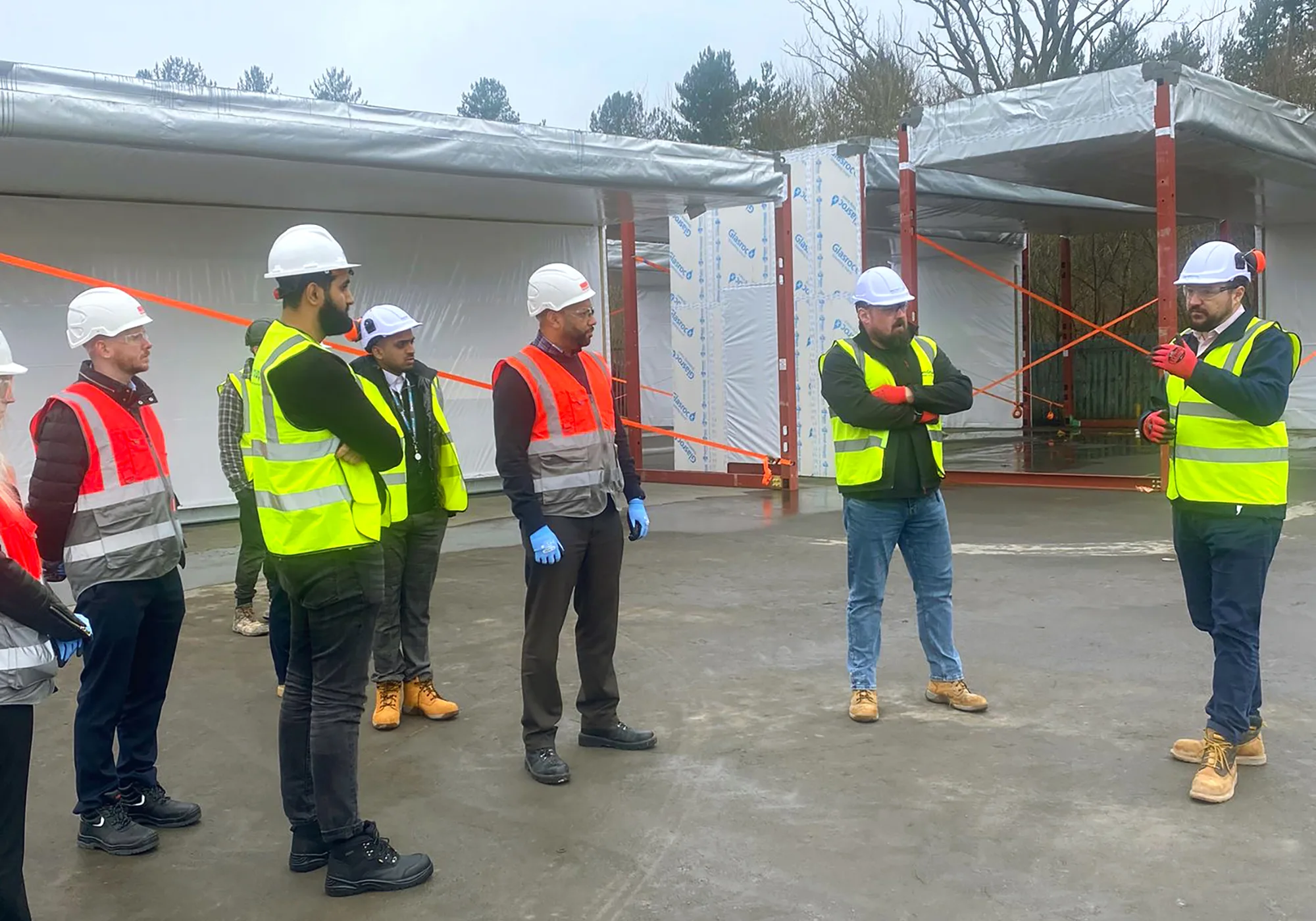 Adrian at the Darwin Group productions facility, showing under construction modules to a group of visitors. All are wearing hi-vis and hard hats.