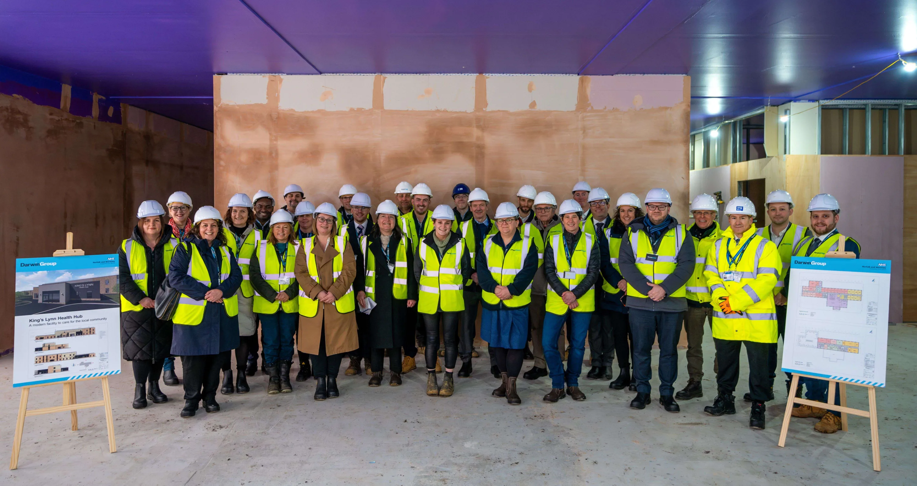 A large group of people in high-vis and hard hats, having just toured the under-construction facility at King's Lynn. The Passive Purple spray coating is still visible on the ceiling and the plaster walls are newly finished.