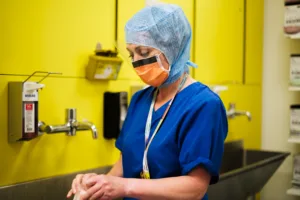 A theatre nurse in full PPE, scrubbing up ready for surgery. Behind them is a large, medical grade sink and a yellow wall.
