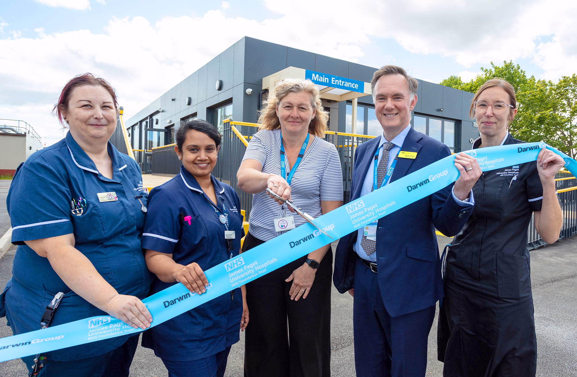 James Paget Orthopaedic Outpatient Centre at Great Yarmouth. Ribbon cutting. A group of clinicians are stood outside with the building in the background. They are holding a blue ribbon and a lady in the middle of the group is holding a large pair of ceremonial scissors.