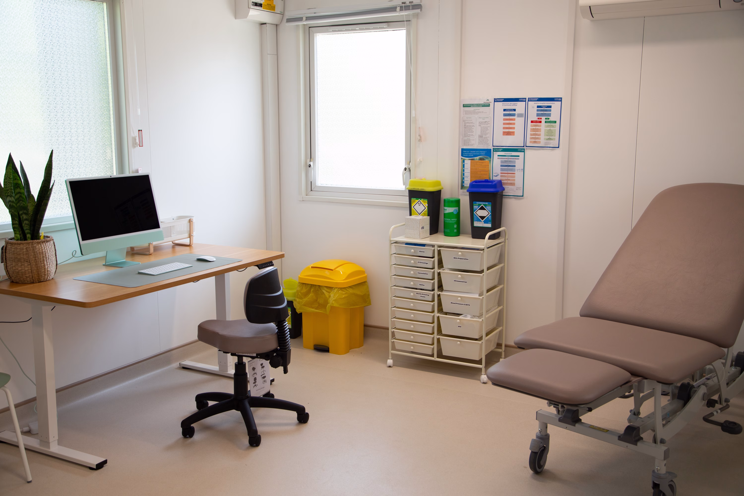 Interior of a healthcare space - consultation room. The floor and walls are light-coloured, There is a desk with a computer, as well as a patient examination bed.