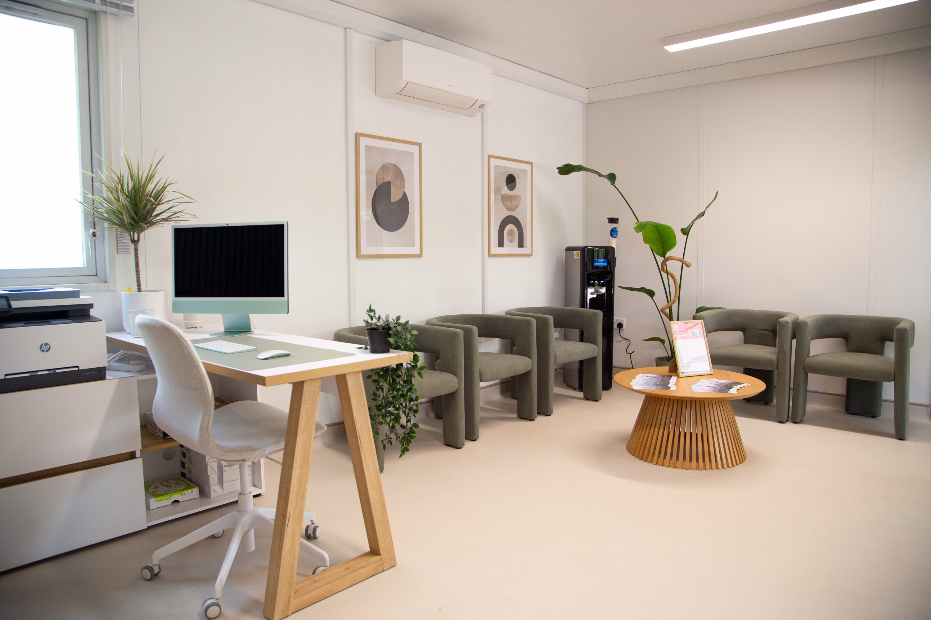 Interior of a healthcare space - waiting/reception area. The floor and walls are light-coloured. There is a wooden desk with a computer on it. In the background are a number of grey chairs and a wooden coffee table.