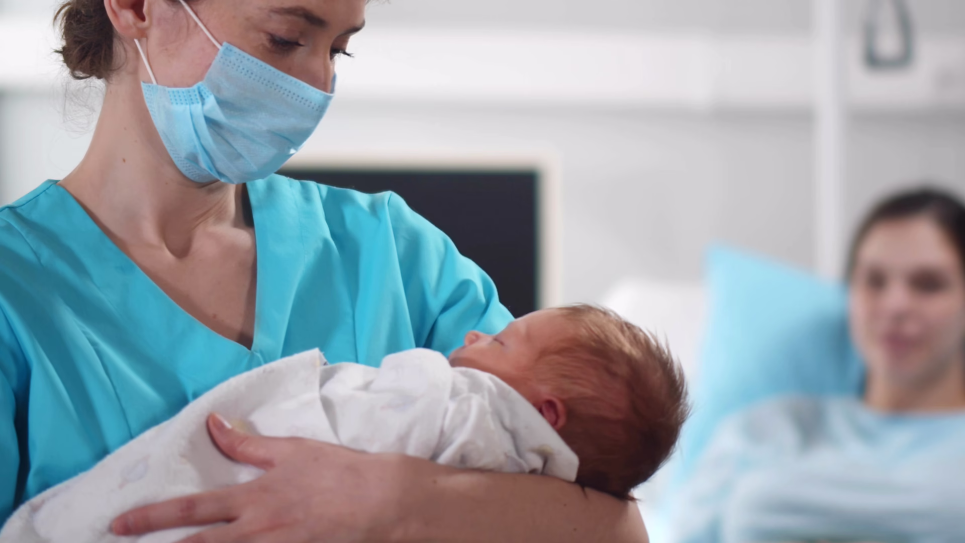 Portrait of nurse wearing safety mask cradling newborn in arms with young mother resting in bed at hospital ward. Happy woman relaxing in bed after childbirth with doctor holding baby