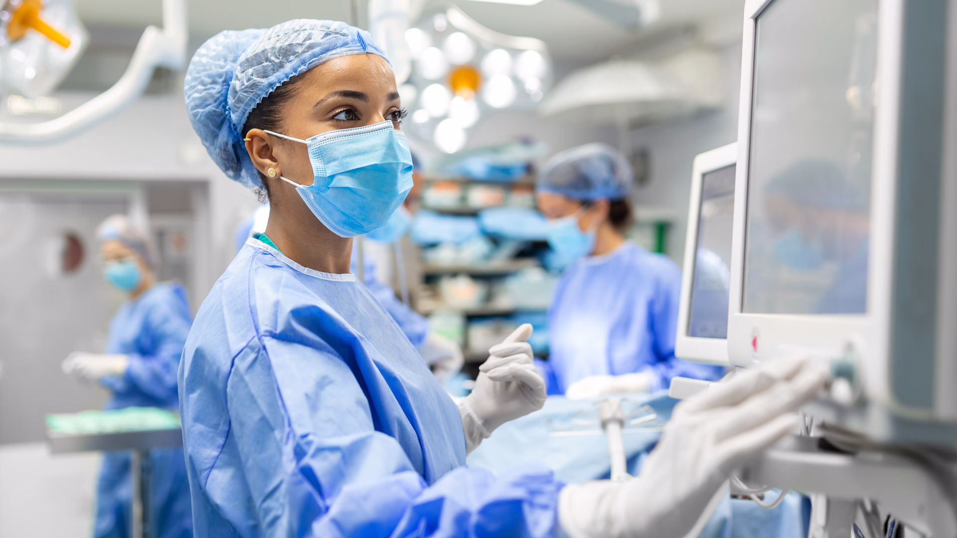 Anesthetist Working In Operating Theatre Wearing Protecive Gear checking monitors while sedating patient before surgical procedure in hospital